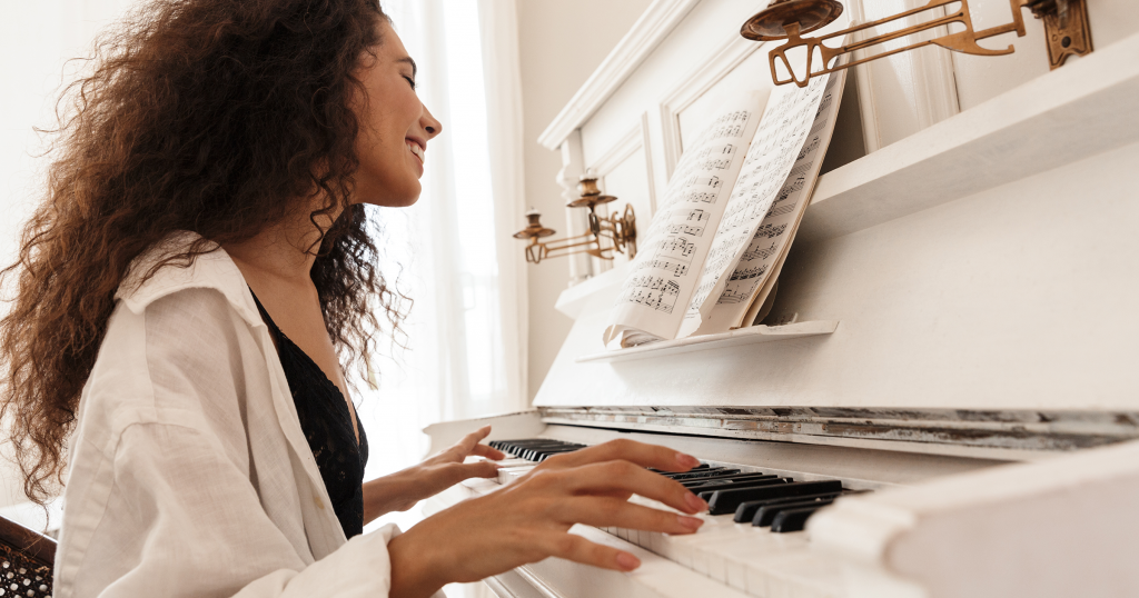 A smiling woman plays a white acoustic cabinet piano while reading sheet music