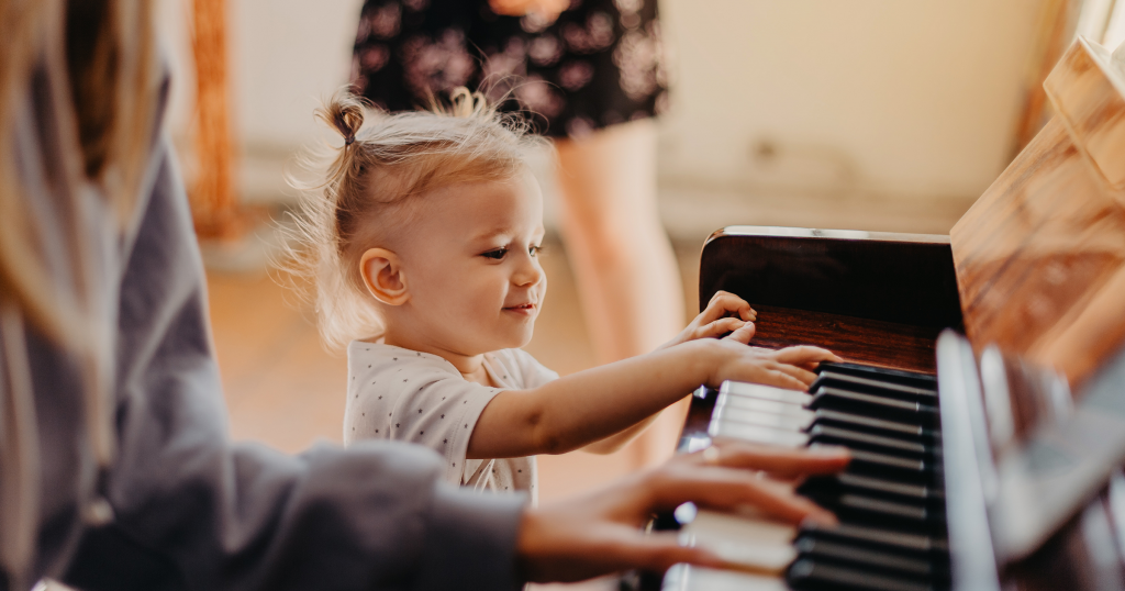 A toddler plays the low register keys of a piano while another person is playing higher up on the keyboard
