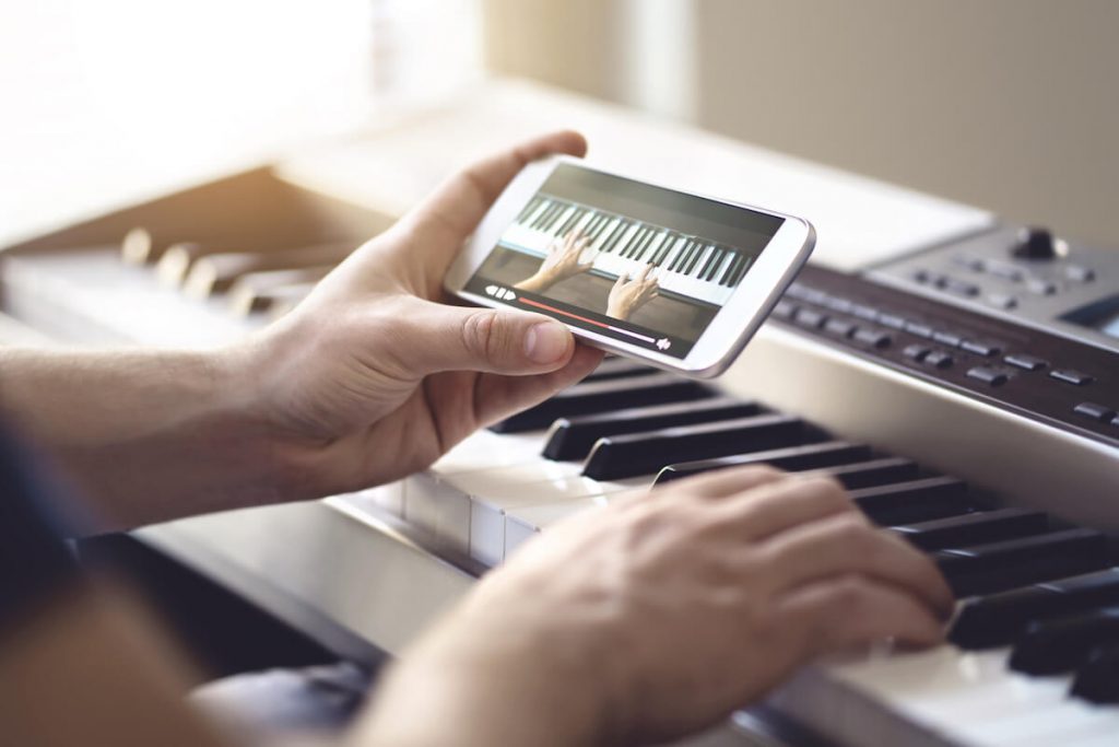 A person holding a phone with their left hand showing a piano tutorial video while their right hand plays on a digital keyboard